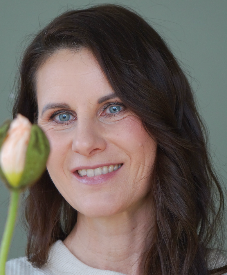 Lächelnde Frau in den Wechseljahren mit blauer Augenfarbe und dunklem Haar, die eine Blüte im Vordergrund betrachtet. Symbol für natürliche Schönheit und Selbstfürsorge in der Lebensmitte